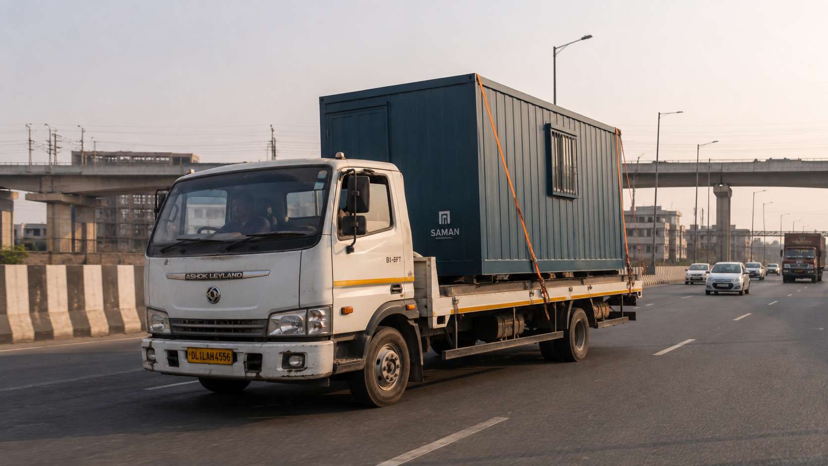Deep Teal porta cabin loaded on a 14-ft truck en route from Greater Noida to a Delhi site — heavy-vehicle window delivery.