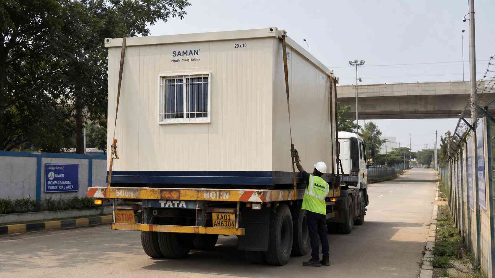 Ivory white SAMAN porta cabin on a flatbed truck mid-dispatch on the Bommasandra Industrial Area internal service road