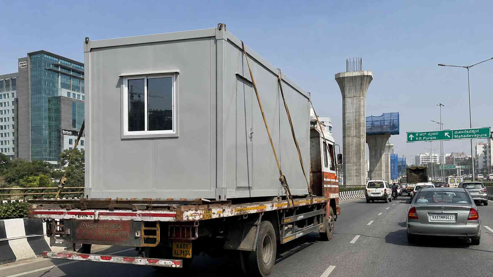 Porta cabin loaded on flatbed truck en route to KR Puram via the Outer Ring Road, Pearl Grey cabin during mid-day delivery window.