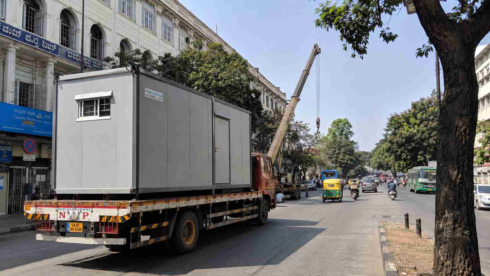 Pearl Grey porta cabin on a flatbed truck on Sampige Road Malleshwaram, with crane ready for offload on the formal-grid commercial stretch.