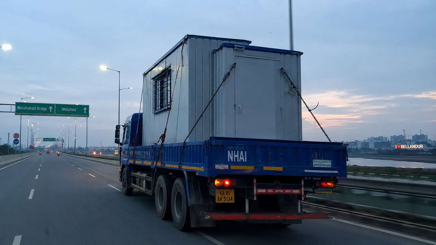 NHAI blue flatbed truck carrying a porta cabin on Bengaluru Outer Ring Road heading toward Marathahalli Bridge.