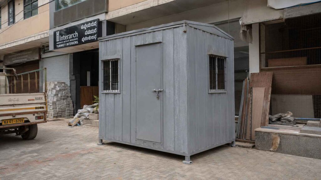 A Steel Grey MS porta cabin parked on a Koramangala 80 Feet Road commercial fitout site at midday.