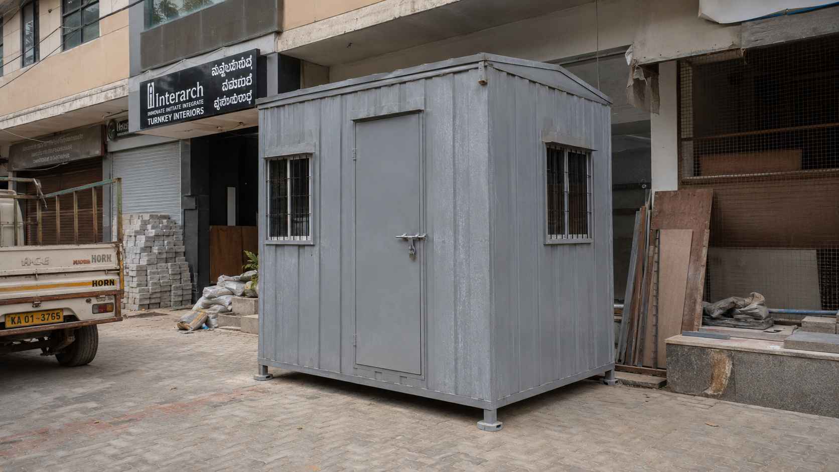 A Steel Grey MS porta cabin parked on a Koramangala 80 Feet Road commercial fitout site at midday.