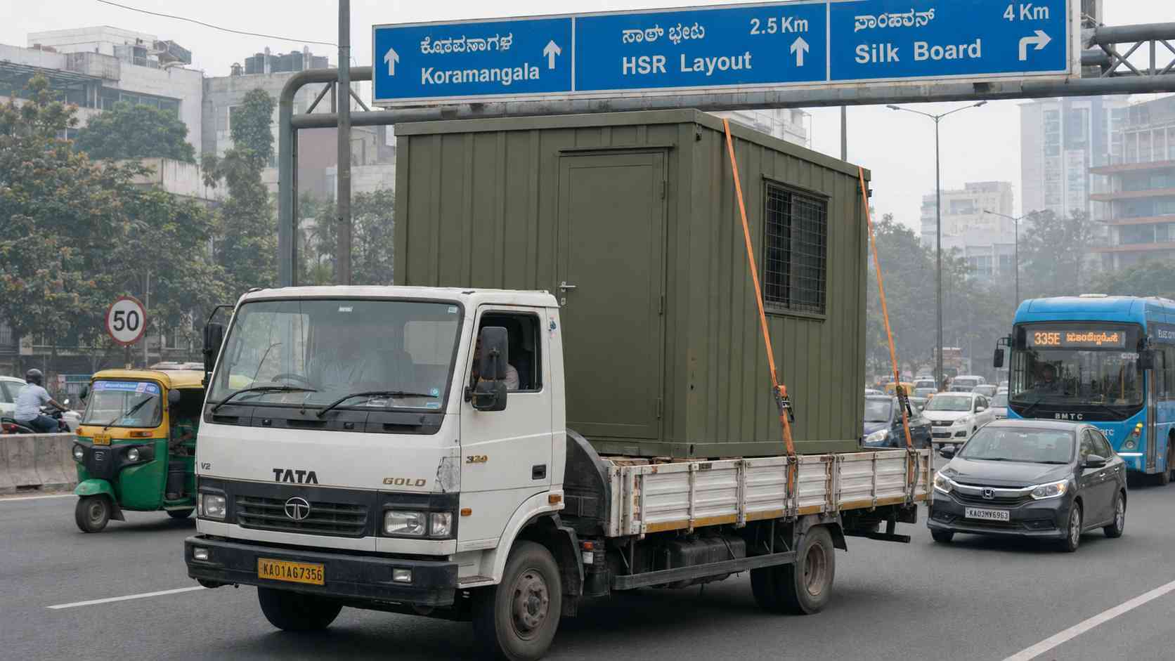  Tata flatbed truck carrying an Olive Drab porta cabin from Gopasandra heading north on Hosur Road towards Koramangala in morning traffic.