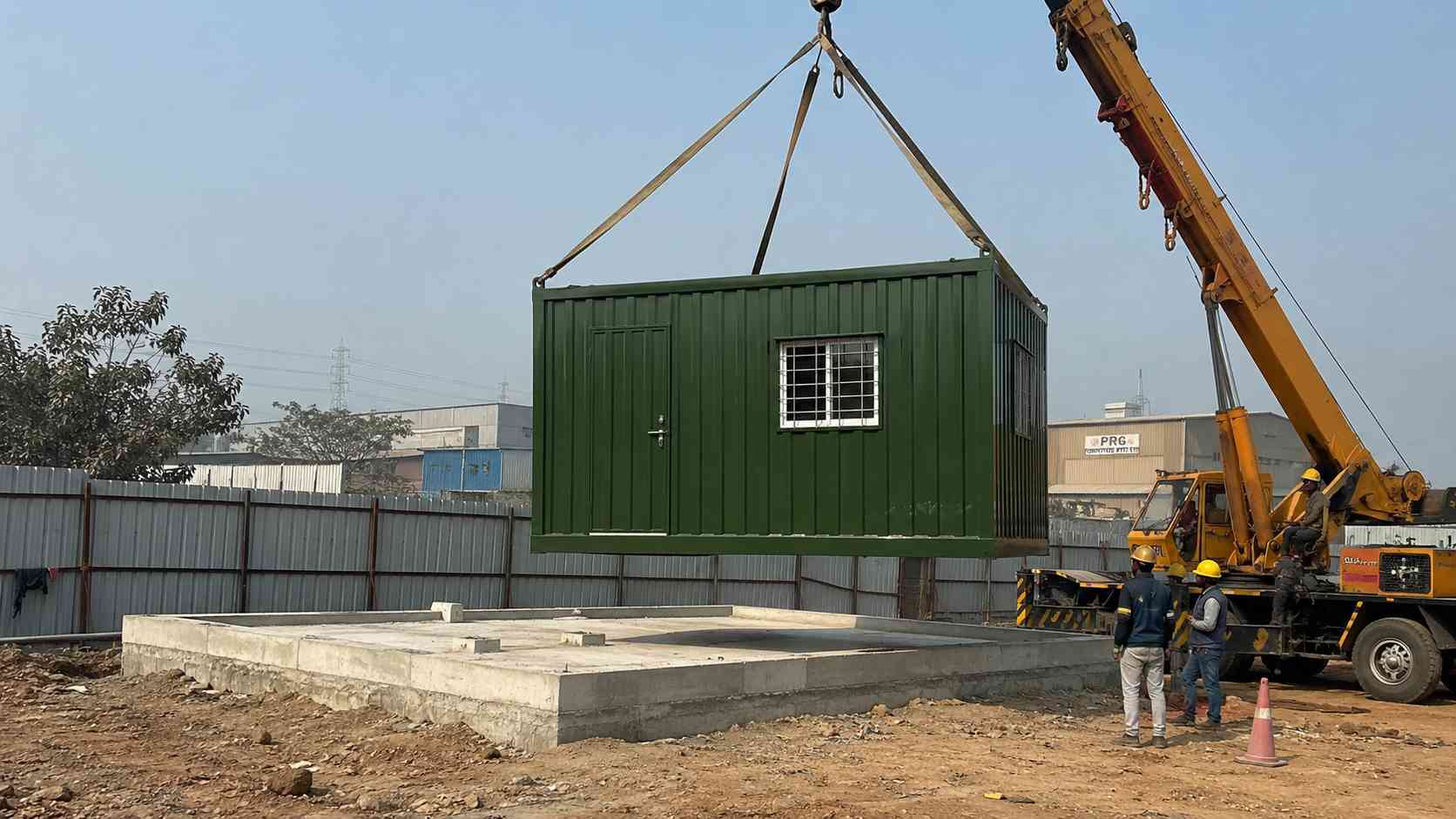 A Forest Green 20×10 ft portable cabin being lowered by crane onto a Delhi industrial cluster site. 