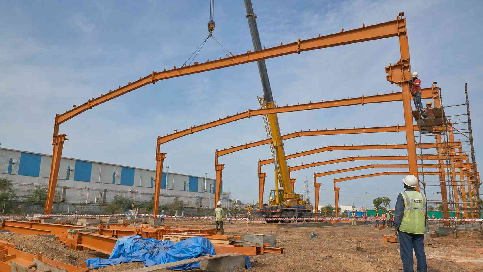 Crew erecting a Safety Orange primed PEB steel frame on site using bolted assembly.