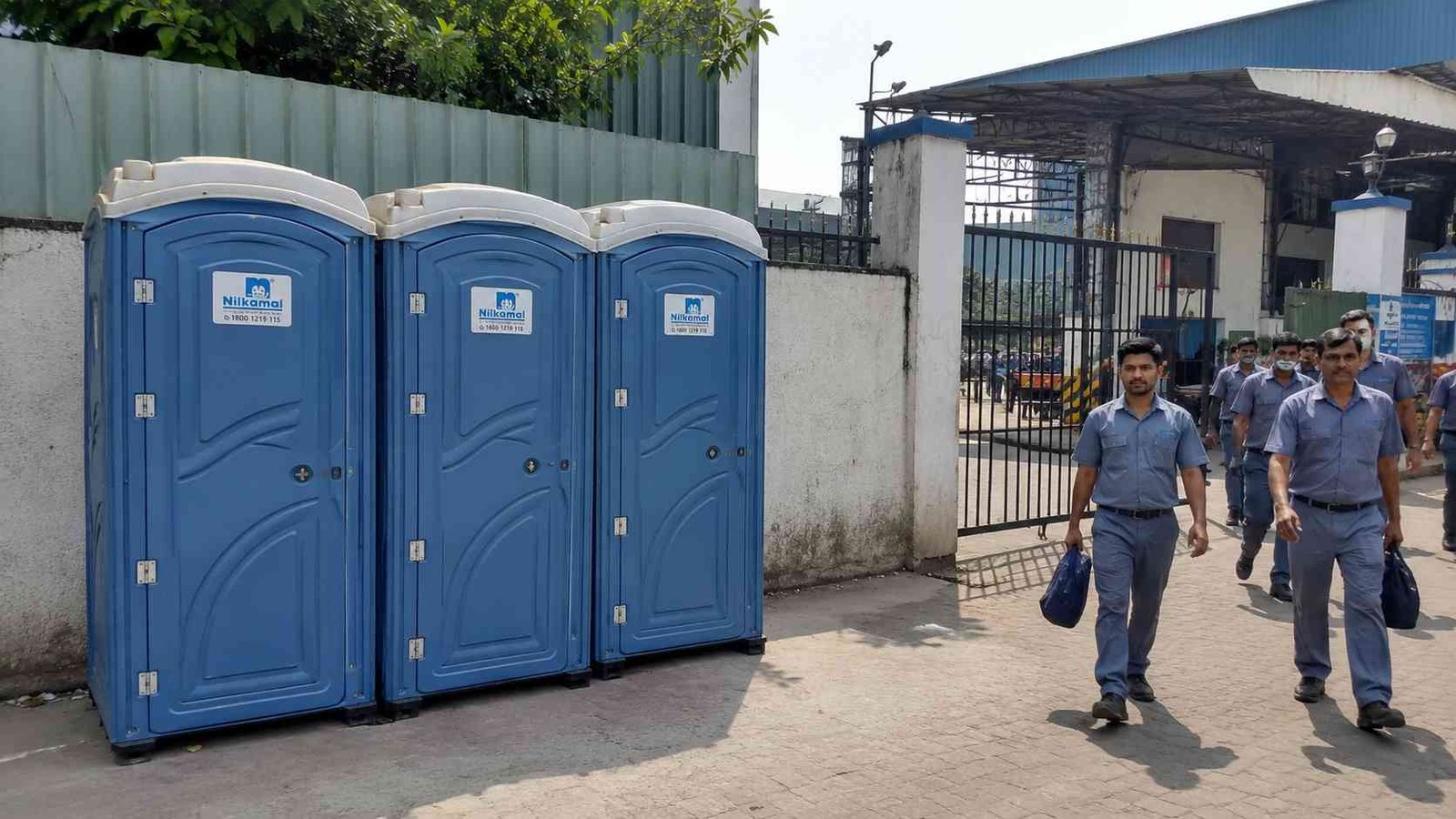 Row of Steel Blue portable toilets at an Indian factory gate during a shift change. 
