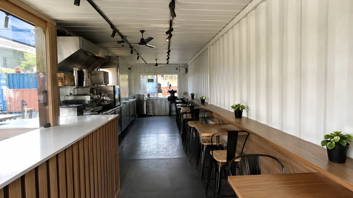 Container cafe interior with counter, compact kitchen and light-wood seating in white insulated unit