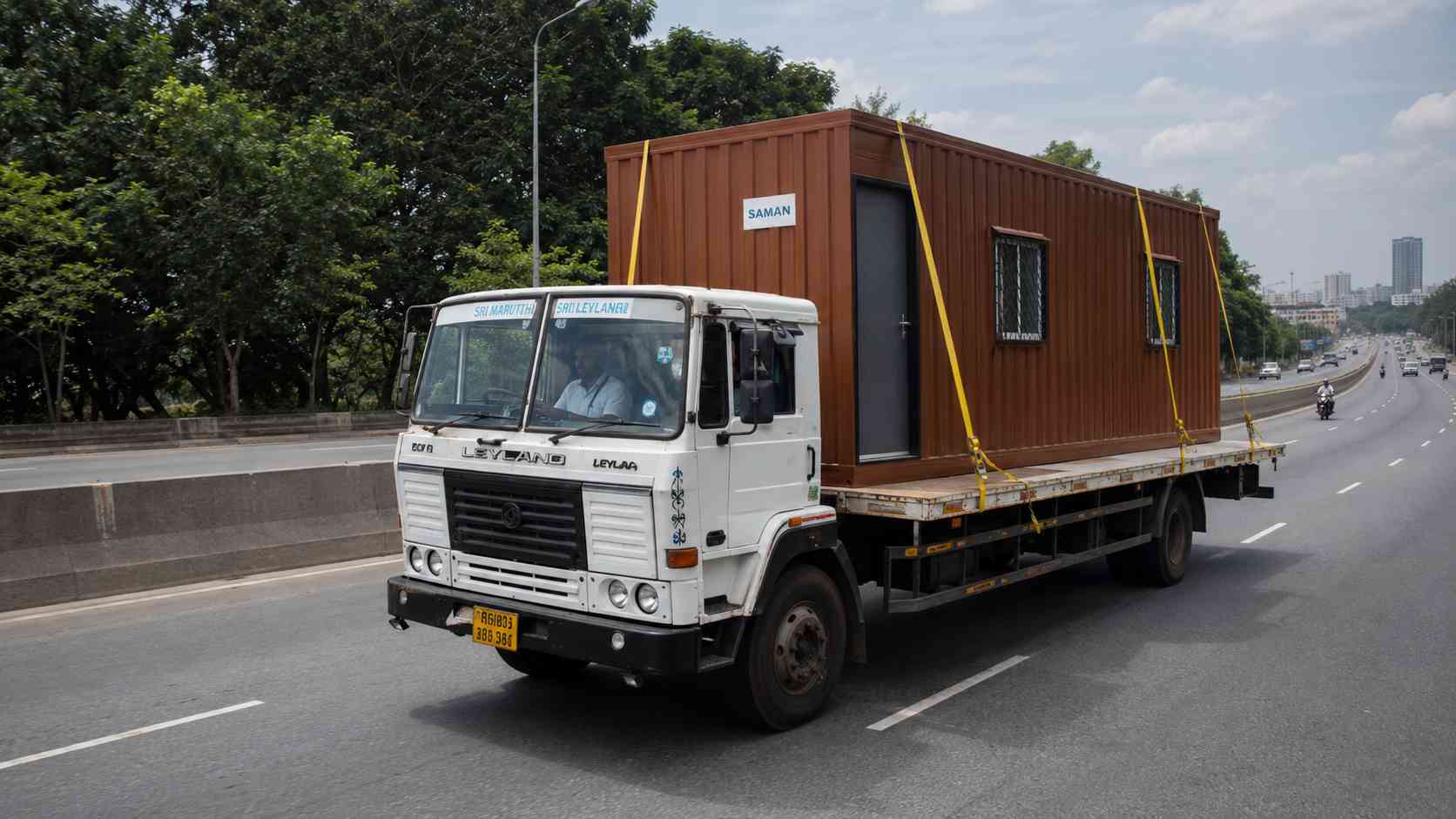 Brick Terracotta SAMAN porta cabin secured on an Ashok Leyland flatbed truck mid-route from Gopasandra to Rajajinagar on the NICE Peripheral Ring Road in Bengaluru.
