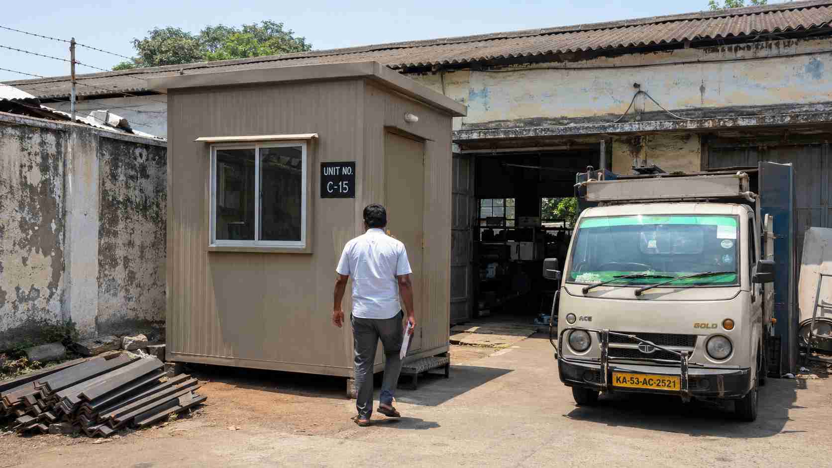 Warm Taupe MS porta cabin used as a supervisor cabin in front of a KSSIDC small-scale unit (Unit C-15) inside Industrial Town Rajajinagar, with a Tata Ace tempo and stacked MS angle iron in the yard.